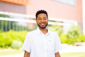 Image of a smiling black man, black hair, white shirt in foreground. Green shrubs and red building in background